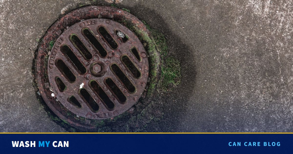A round rusty storm drain grate set in gray concrete, with a dark water stain and green moss around its edge