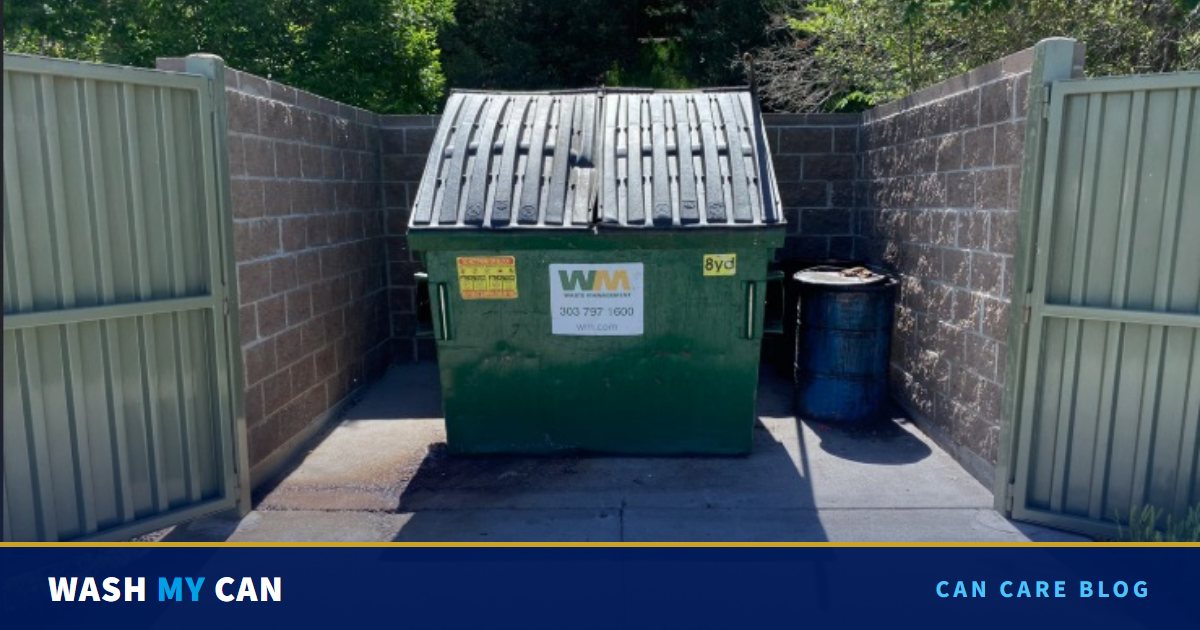 A green front-load dumpster inside a block wall enclosure with open metal gates, with a dark stain spreading across the concrete pad in front of it