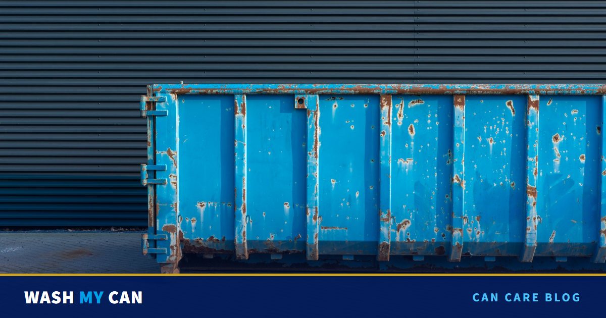A rusty blue industrial waste container parked on a brick paved lot against a dark corrugated metal wall
