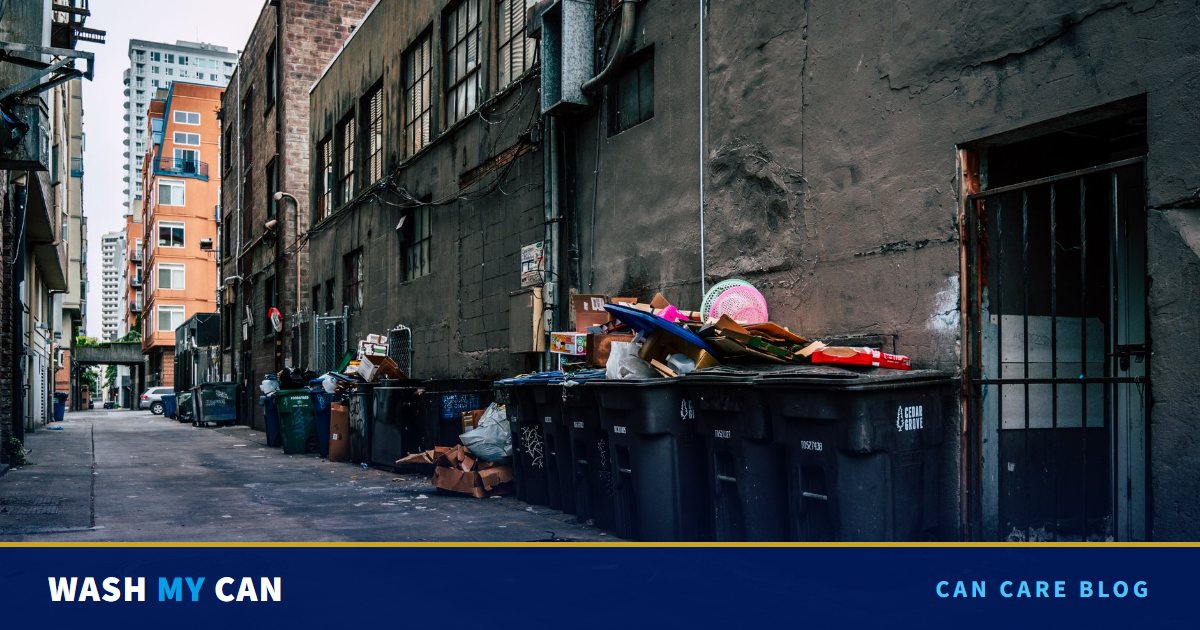 A city back alley lined with black wheeled trash carts, several overflowing with boxes and bags, beside a gritty building wall