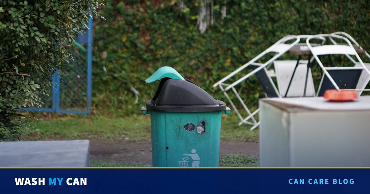 A weathered green trash can with a black lid sitting alone on a patch of grass beside a hedge