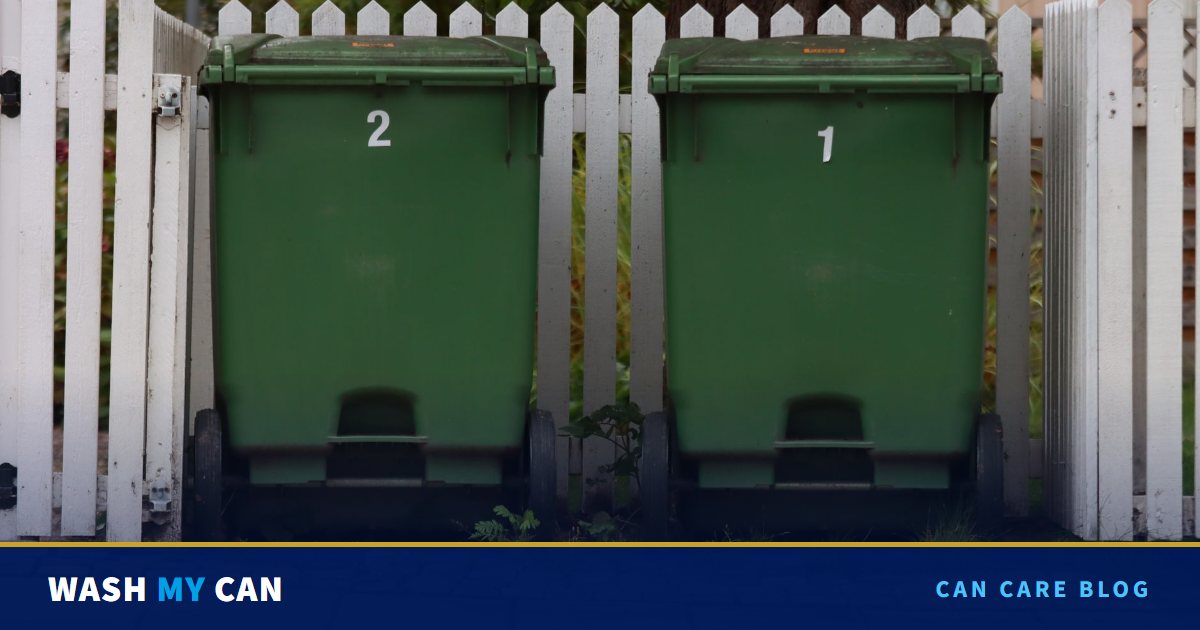 Two green wheelie trash cans lined up against a white picket fence in front of a home